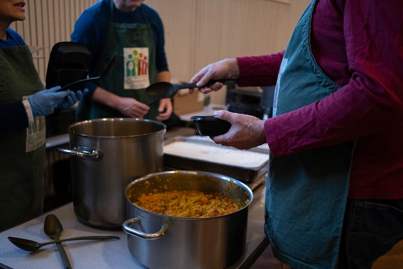 A group of volunteers wearing Community Kitchen aprons scoop a pot of red food into containers